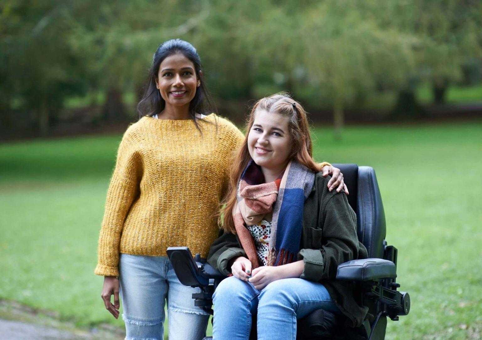 Two friends smiling outdoors, one using a wheelchair